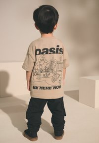 Beige bomulds t-shirt med sort trykt kunstværk, der viser en scene; parret med sorte cargo bukser. Teksten lyder "oasis" og "BE HERE NOW."