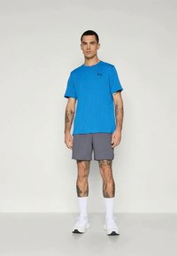 Blaues T-Shirt mit kurzen Ärmeln, strukturiert und mit einem subtilen Muster, kombiniert mit grauen Shorts. Das Model trägt weiße Sportschuhe und Knöchelsocken.