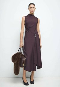 Bordowa sukienka bez rękawów z dopasowanym gorsetem i drapowanym przodem. Do tego brązowa futrzana torebka i czarne buty na wysokim obcasie.