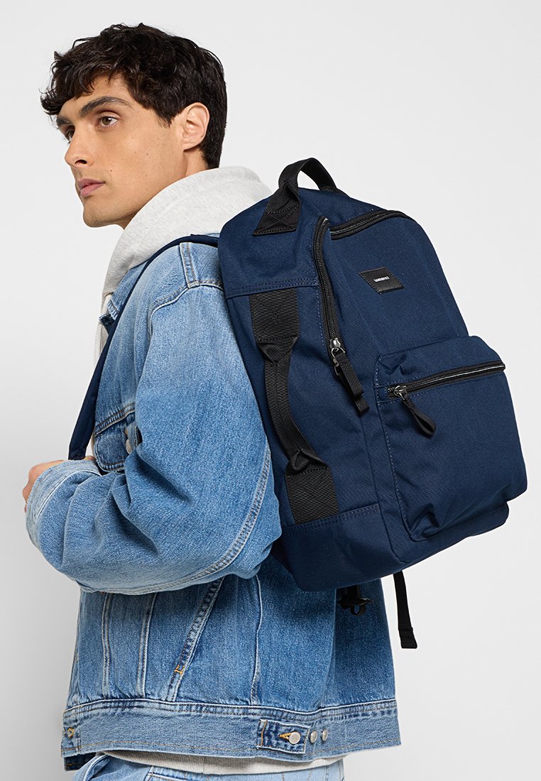 Mochila azul marino con dos bolsillos delanteros con cremallera, correas ajustables y un material texturizado. Lleva sobre el hombro contra una chaqueta de mezclilla.