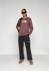 Γυναίκα με σκούρα γυαλιά ηλίου, μωβ φούτερ The North Face, μαύρο παντελόνι cargo, ροζ αθλητικά και κρατώντας μια μαύρη ζώνη σακιδίου.