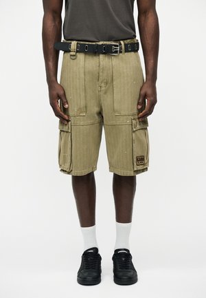 Mand iført olivengrønne cargoshorts med flere lommer, et sort bælte, hvide sokker og sorte sneakers, stående mod en hvid baggrund.