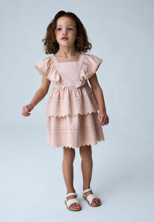 Junges Mädchen mit lockigem Haar, das ein hellrosa Kleid mit Rüschenärmeln und weiße Sandalen trägt, steht vor einem schlichten Hintergrund.