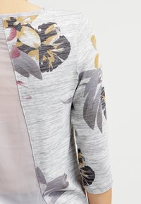 Cardigan floreale grigio con maniche 3/4, tessuto grigio chiaro dalla texture leggera, grandi motivi floreali neri e dorati e un pannello posteriore trasparente.