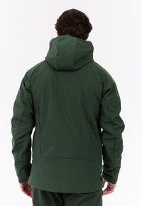 Giacca impermeabile verde con cappuccio, caratterizzata da una texture liscia e un design aderente. Include tasche laterali e un dettaglio del logo vicino all'orlo.