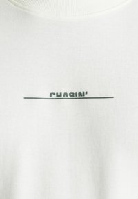 Felpa bianca in cotone con un sottile logo "CHASIN'" grigio e una linea orizzontale, dotata di collo a coste e texture liscia.