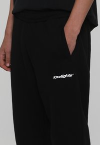 Černé tepláky s elastickým páskem, bočními kapsami a bílým logem "lowlights" potištěným na stehně. Hladká textura látky.