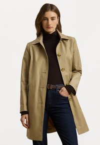 Hnedý trench coat s golierom a zapínaním na gombíky, vyrobený z hladkej tkaniny. Nosený cez tmavý rolák a skombinovaný s tmavými džínami.