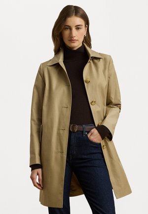 Cappotto beige con colletto e chiusura a bottoni, realizzato in un tessuto morbido. Indossato sopra un dolcevita scuro e abbinato a jeans scuri.