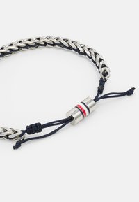 Silverflätad armband med mörkblått snöre och en metallklämma som har röda och blå accenter, säkrad med justerbara knutar.