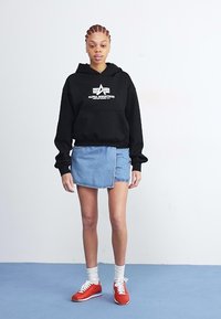 Čierna mikina s kapucňou a bielym logom, nosená s modrou džínsovou sukňou. Model má tiež na sebe červené tenisky a biele ponožky na modrej podlahe.