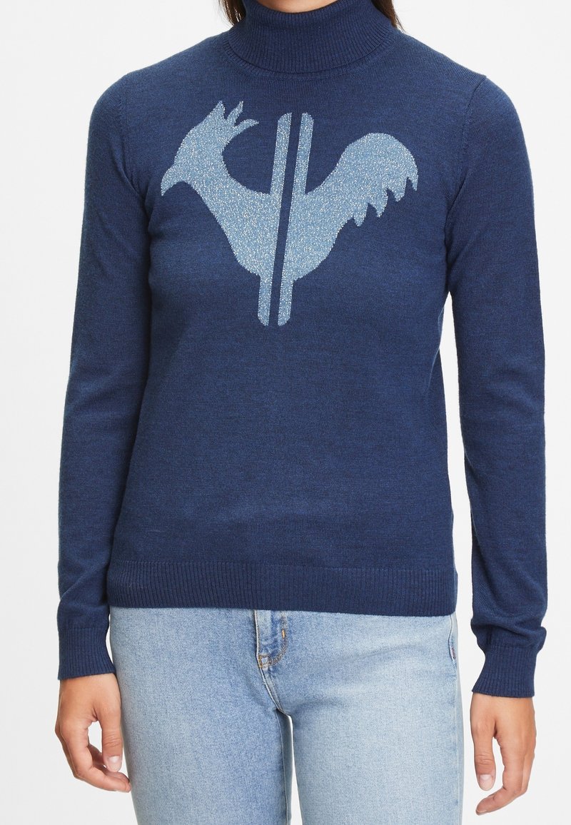 Femme portant un pull à col roulé bleu marine avec un motif stylisé de coq bleu clair et un jean bleu clair.