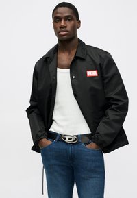 Μαύρο ελαφρύ μπουφάν με κόκκινο λογότυπο "DIESEL", με κούμπωμα σναπ και ελαστικές μανσέτες, φορεμένο πάνω από λευκό tank top και μπλε τζιν.