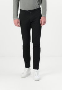 Svarta slim-fit byxor med en slät textur, utrustade med knappstängning och dragkedja. Kombineras med vita sneakers, lämpliga för avslappnad användning.