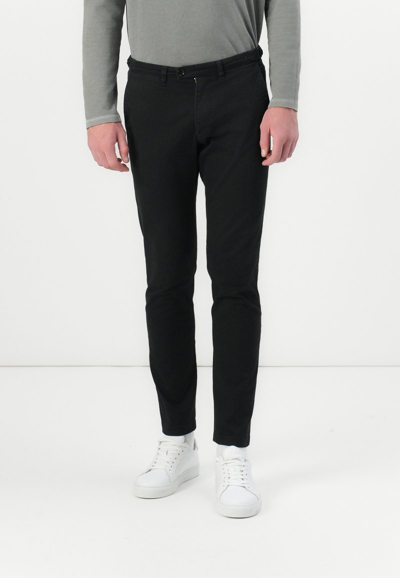 Svarta slim-fit byxor med en slät textur, utrustade med knappstängning och dragkedja. Kombineras med vita sneakers, lämpliga för avslappnad användning.