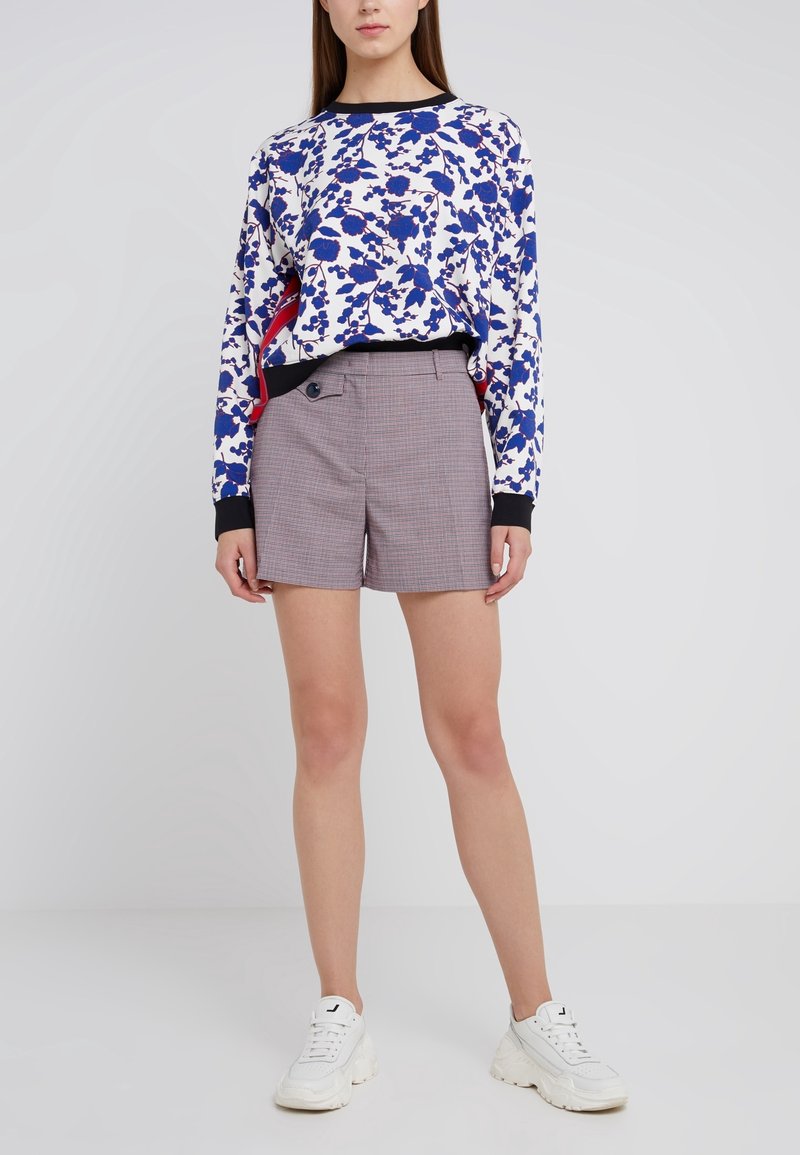 Femme portant des baskets blanches, un short à carreaux avec des détails à boutons, et un haut à manches longues blanc avec des accents floraux bleus et rouges.