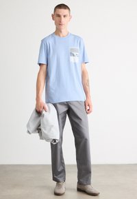 Lyseblå bomull T-skjorte med lomme og grafisk design, kombinert med grå bukser og beige sneakers. Personen holder en grå jakke.