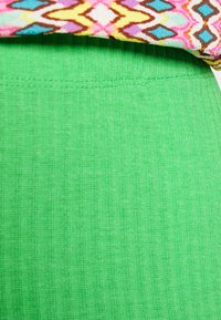 Leggings verdes com nervuras apresentam um tecido texturizado e uma cintura ajustada. Uma blusa com padrão é visível na parte superior, adicionando contraste.