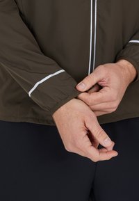 Veste de sport marron avec des accents blancs, dotée de poignets élastiques. Le matériau semble léger et lisse, avec une fermeture éclair.