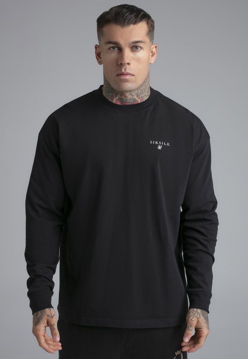 Mladý muž s tetováním a propíchnutím nosí černé tričko s dlouhým rukávem značky SikSilk, stojící před jednobarevným šedým pozadím.