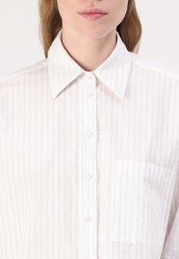 Camicia bianca con bottoni, a righe verticali rosa, dotata di colletto classico, taschino frontale e tessuto leggermente strutturato.