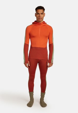 Man staat rechtop en draagt een oranje en roestkleurige lange mouwen hoodie met bijpassende leggings met groene zijstroken, en dikke gebreide sokken.