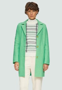 Manteau vert menthe avec un col en V et trois boutons, porté sur un pull rayé, assorti à un pantalon crème. Texture douce et coupe ajustée.