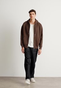 Brown hooded sweatshirt with front pockets, worn over a white ribbed t-shirt, paired with dark jeans and white sneakers. Smooth texture.