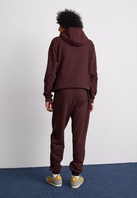 Hnědá mikina s kapucí a odpovídající tepláky vyrobené z měkké látky. Obsahuje kapuci na stahování a žebrované manžety. Žluté a šedé tenisky dokončují celý outfit.