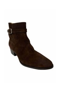 Botín de ante marrón hasta el tobillo con punta afilada y una correa con hebilla plateada. Tacón bajo y textura suave. Diseño sencillo.