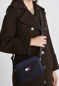 Mörkblå crossbody-väska med ett meshnät på framsidan, prydd med en Tommy Jeans-logotyp och en svart rem med vita detaljer. Buren över en brun kappa.
