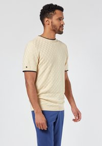 Suéter de punto beige con un patrón de cuadros texturizado, mangas cortas, cuello y dobladillo de canalé negro, combinado con pantalones azules.