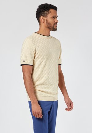 Suéter de punto beige con un patrón de cuadros texturizado, mangas cortas, cuello y dobladillo de canalé negro, combinado con pantalones azules.