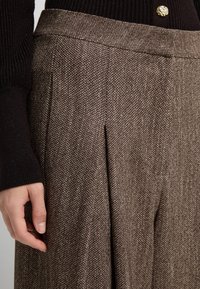 Pantaloni a scacchi marroni realizzati in tessuto strutturato, con pinces e una cintura liscia, abbinati a un top scuro a costine.