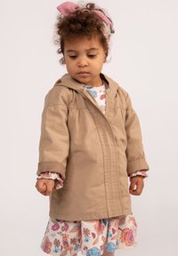 Jasnobrązowa lekka kurtka z kapturem, z zamkiem błyskawicznym z przodu i detalami w postaci szwów, noszona na kwiecistej sukience z falbanami na rękawach.