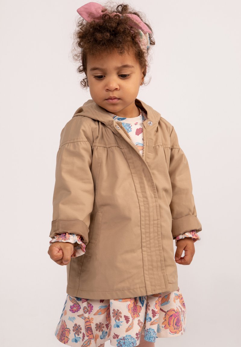Jasnobrązowa lekka kurtka z kapturem, z zamkiem błyskawicznym z przodu i detalami w postaci szwów, noszona na kwiecistej sukience z falbanami na rękawach.