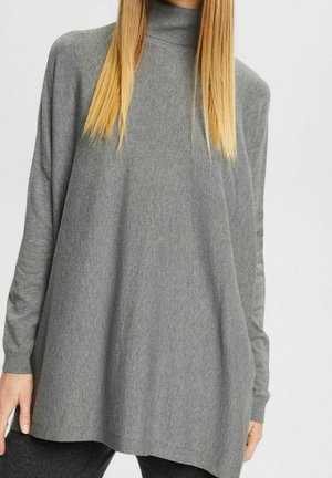 Femme portant un pull gris ample à col roulé et manches longues, avec des cheveux blonds raides visibles sur les épaules.