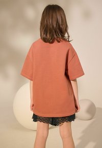 Brændt orange oversize t-shirt med korte ærmer, parret med en sort nederdel med blondekant, set fra ryggen. Glat stof tekstur.