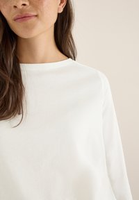 Haut à manches longues blanc avec un col rond. Tissu en coton lisse, montré partiellement, avec une coupe légèrement décontractée et une texture naturelle.