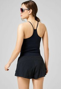 Sort atletisk tanktop med tynde stropper parret med sorte shorts med bølget kant og perforerede detaljer. Model iført solbriller.