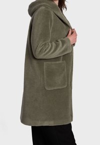 Cappotto in pile verde oliva con cappuccio, caratterizzato da una vestibilità comoda, ampia tasca laterale e tessuto testurizzato per il calore.