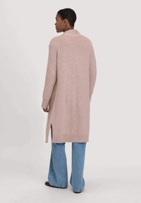 Persoon in lange beige cardigan met zijsplitten over blauwe wijde jeans en zwarte schoenen, staande tegen een effen lichte achtergrond.