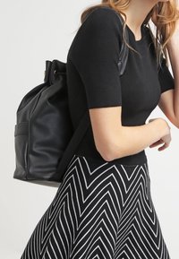 Zwarte leren rugzak met een trekkoordsluiting en één voorvak, gedragen over een strakke zwarte top en een rok met zigzagpatroon.