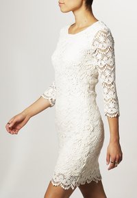 Vit spetsklänning med trekvartsärmar, med intrikata mönster och en scallopad nederkant, figurnära design och texturerad tyg.