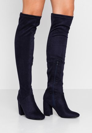 Granatowe zamszowe kozaki za kolano z grubym obcasem i zamkami po bokach, noszone na gołych nogach na białym tle.