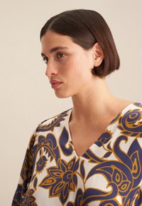 Weiße V-Ausschnitt-Bluse mit einem blau-goldenen Blumenmuster. Glatte Stoffqualität mit leichtem Glanz und lockerer Passform.