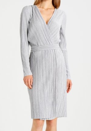 Kobieta ubrana w jasnoszarą, długorękawową plisowaną sukienkę kopertową z dekoltem w kształcie litery V i dopasowanym paskiem w talii, stojąca na jednolitym tle.