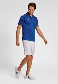 Blå polo skjorte med korte ærmer, hvide detaljer og logo; parret med hvide shorts og hvide sneakers med sorte striber.