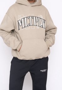Felpa beige con cappuccio e scritta nera "MULTIPLY", tasca frontale, polsini a coste, abbinata a pantaloni jogger neri con logo bianco.
