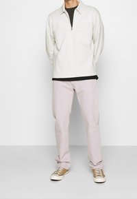 Hombre vistiendo un suéter de medio cierre color blanco roto, camiseta negra debajo, jeans rosa claro y zapatillas color beige, de pie contra un fondo blanco liso.
